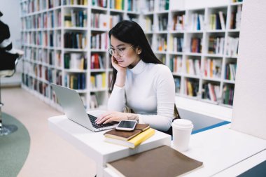 Sıkıcı Asyalı yetişkin gözlüklü ve zarif giysileri olan bir öğrenci elinde not defteriyle masada otururken dizüstü bilgisayarında daktilo yazıyor ve içkileri götürüyor.