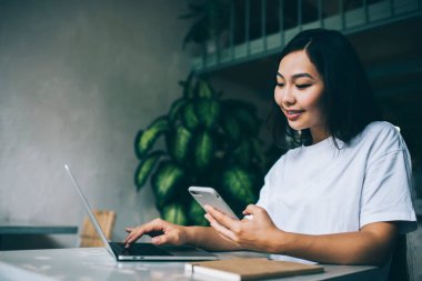 Gülümseyen, siyah saçlı, açık kafede oturmuş bilgisayarını karıştıran ve akıllı telefondan mesaj atan serbest bir etnik kadın.