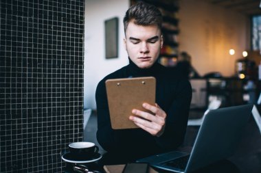 Konsantre olmuş erkek serbest çalışan rahat bir iş yerinde dizüstü bilgisayar kullanarak masada oturuyor ve uzaktan çalışırken panoyu tutuyor ve kahve içiyor.