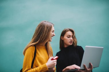 Modern dizüstü bilgisayarda gündelik kıyafetlerle gezen konsantre kız öğrenci turkuaz duvarın yanında mutlu bir arkadaşla vakit geçiriyor.