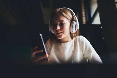 Gündelik giysiler içinde gülümseyen kadın ve modern kulaklıklar akşam saatlerinde cep telefonuyla video görüşmesi yapıyor.