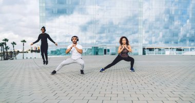 Aktif giyimli genç atletik arkadaşlar yarışmadan önce dışarıda antrenman yaparken şehir meydanında modern cam bina yakınında fiziksel egzersiz yapıyorlar.