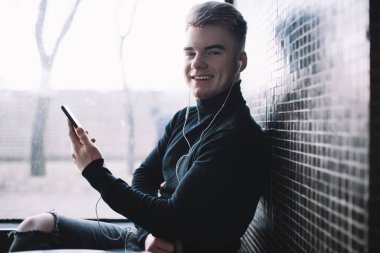 Siyah balıkçı yakalı ve yırtık pantolonlu neşeli genç adam pencerenin yanındaki duvara yaslanırken parlak bir gülümsemeyle kameraya bakıyor.