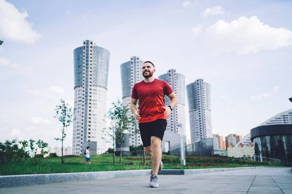Full body of cheerful male runner in sportswear and sneakers jogging on pavement during training in summer city and looking away