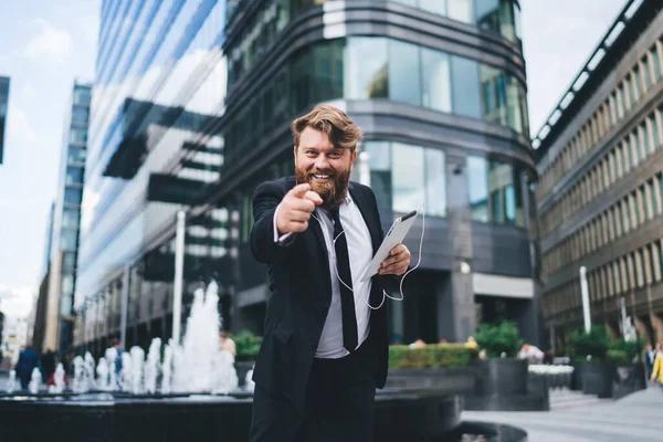 Happy entrepreneur in business suit with tablet and earphones pointing at camera standing on street near contemporary buildings and fountain