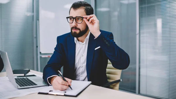 Hombre Concentrado Traje Formal Ajustando Gafas Escribiendo Portapapeles Mientras Está —  Fotos de Stock