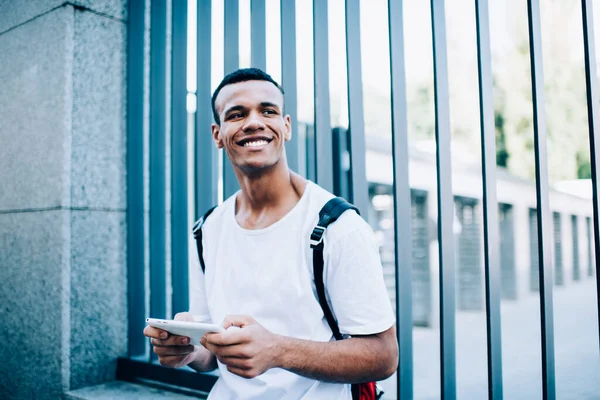 Smiling African American male looking away and browsing cellphone while standing near metal enclosure with pillar on street in city