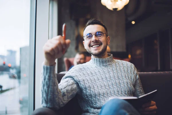 Happy bearded man in casual outfit and eyeglasses smiling and looking away while writing thoughts in notepad in cozy cafe