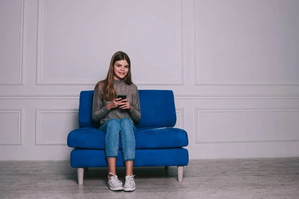 Full body of happy in casual clothes with mobile phone smiling and looking at camera while sitting on sofa against white wall