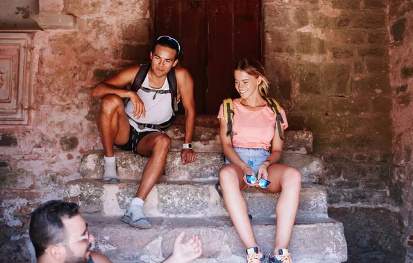 Cheerful young woman and men in casual clothes with backpacks resting on stone steps and talking happily while backpacking together