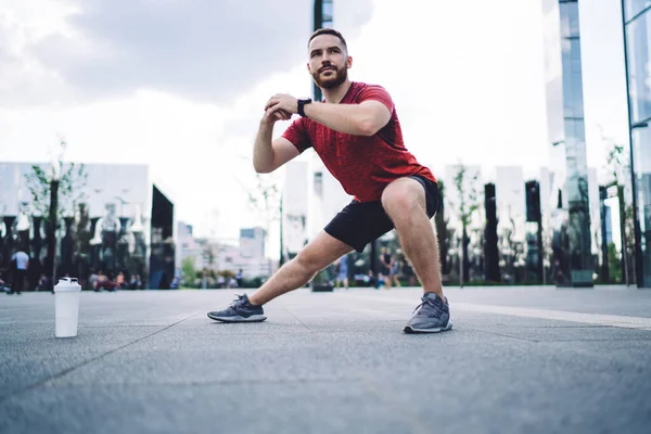 Full body of concentrated bearded sportsman in activewear and sneakers standing on paved street near plastic bottle of water and stretching legs before workout