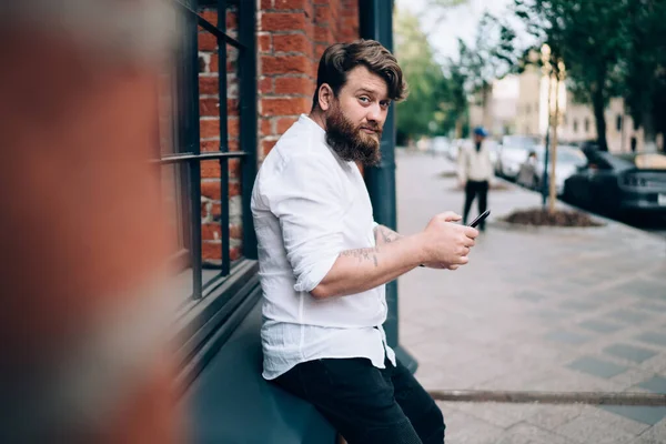 Pencere kenarına yaslanmış sakallı bir adamın kaldırımda dikilip, cep telefonuyla kameraya bakarken yan görüntüsü.