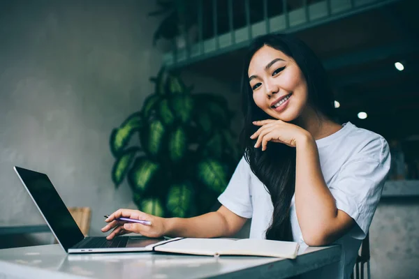 身穿休闲装 面带微笑的亚裔女自由撰稿人坐在桌旁 看着相机触摸下巴 浏览笔记本电脑 — 图库照片