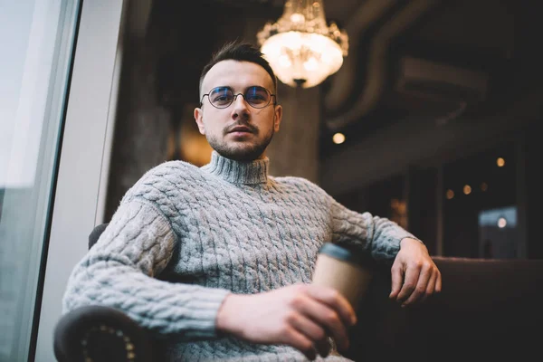 Düşük açılı kendine güvenen sakallı bir erkek. Günlük kıyafet ve göz giysisi. Bir fincan kahve içip kameraya bakıyor.