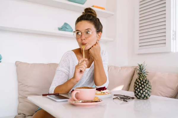 Positive female in casual wear and glasses enjoying eating healthy breakfast while looking away dreamily during summer vacation on Bali
