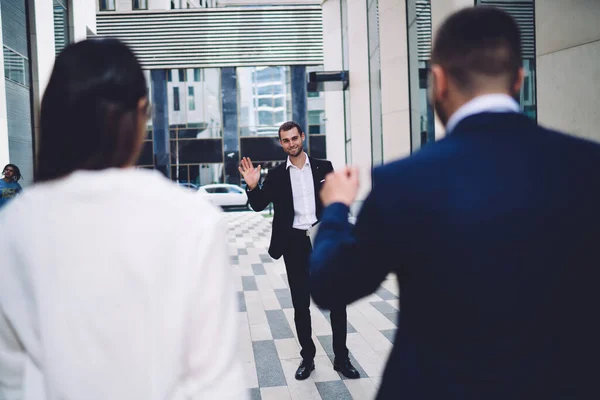 Young elegant office worker in formal wear smiling and waving to colleagues waiting for him near modern glass business center