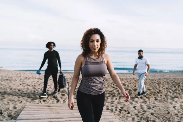 Half length portrait of curly female fitness coach with perfect figure walking at seashore and looking at camera during workout meeting with blurred