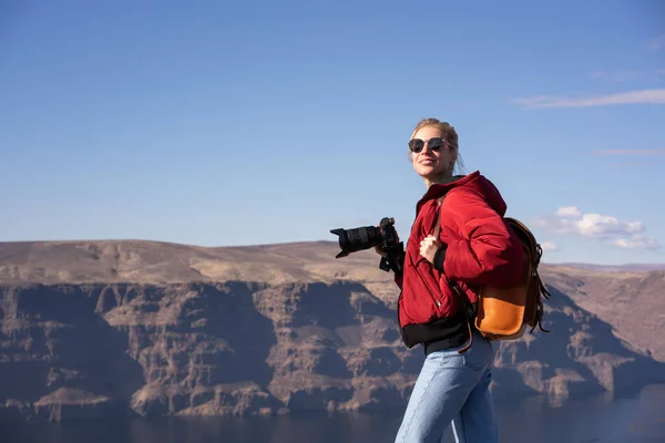 Side view of female explorer in sunglasses with backpack and photo camera looking away while enjoying vacation on nature with wonderful natural landscape from