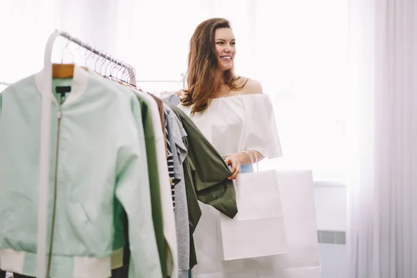 白い紙のショッピングバッグと白いドレスの幸せな女性のサイドビュー笑顔と離れて現代の光の店でジャケットを選択しながら見て — ストック写真