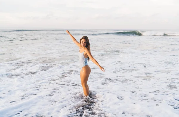 サイドビューの完全な長さ正のスリム女性で白い水着姿で立って腕が泡の海の波によって洗浄海岸に延長 — ストック写真