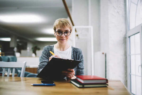 Glad female in casual clothes with eyeglasses and short hair looking at camera sitting at table with clipboard writing report in office