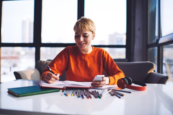 Cheerful female student in casual outfit with short hair taking notes in notepad and working with smartphone against blurred window in workplace