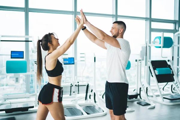 Side view of content sportswoman in active wear with smiling partner giving five against fitness machines while looking at each other