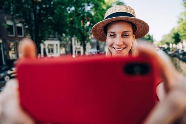 Smiling female blogger with blond hair taking selfie on cellphone while standing near house on embankment during daytime in Amsterdam