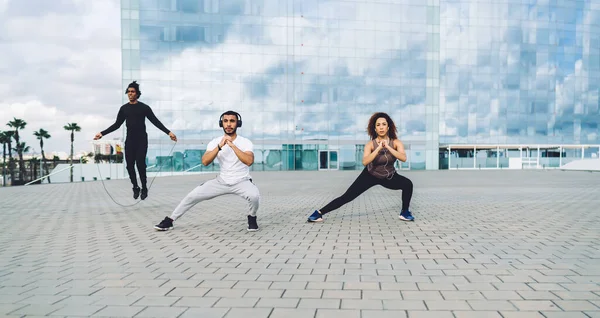 Aktif giyimli genç atletik arkadaşlar yarışmadan önce dışarıda antrenman yaparken şehir meydanında modern cam bina yakınında fiziksel egzersiz yapıyorlar.