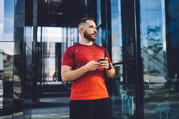 Calm male in sport wear standing near office entrance and checking notifications on smartphone while looking away during daytime in city