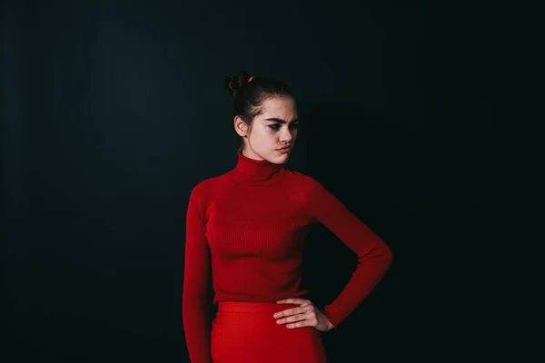 Young female model in red outfit thoughtfully looking away and making faces while standing on black background and holding hand on waist