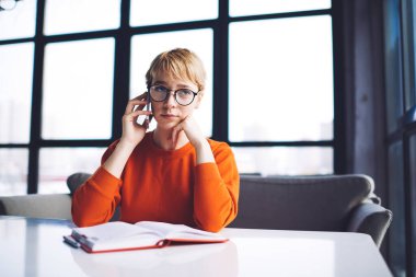 Uzaktan kumandalı, kısa saçlı, dalgın bir kadın. Masaya yaslanmış, iş sırasında telefonla konuşuyor.