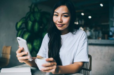 Genç Asyalı kadın kameraya bakıyor ve akıllı telefondan mesaj yolluyor. Kahve içerken molada arka planda bulanık kahve içiyor.
