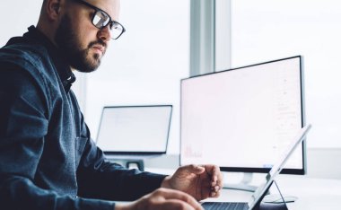 Gözlüklü genç erkek komisyoncunun masaüstü bilgisayarının yanında tablet ekranı izlerken grafikle işteki verileri analiz ederken yan görünümü