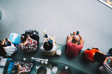 Havaalanındaki bankta oturan ve uçuşunu beklerken akıllı telefonları kullanan çok ırklı isimsiz bir grubun en üst görüntüsü.