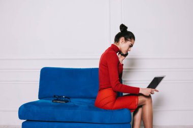 Kırmızı elbiseli, cep telefonuyla konuşan, dizüstü bilgisayarda sörf yapan mavi koltukta oturan genç bir kadının yan görüntüsü.