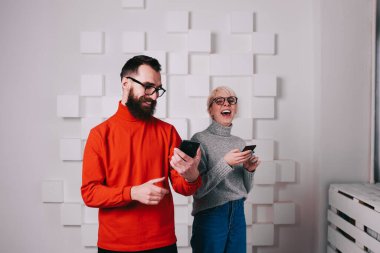 Genç mutlu adam ve kadın, beyaz duvarın yanındaki akıllı telefondaki komik fotoğraflara gülüyorlar.