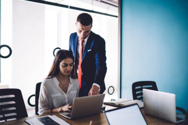 Seçkin takım elbiseli kendine güvenen genç erkek yönetici, ofiste çalışan etnik kadın meslektaşın yanında dururken dizüstü bilgisayarın ekranını işaret ediyor.