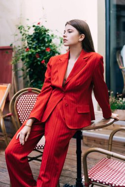 Uzun saçlı, şık kıyafetli, kendine güvenen bir kadın. Açık kafede masaya yaslanıyor ve rüya gibi bir şekilde başka tarafa bakıyor.