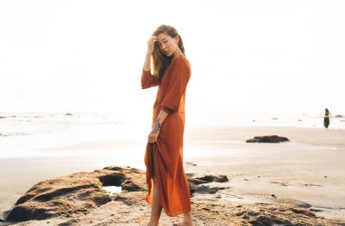 Bali 'de kumlu sahilde dalgalanan okyanusun kıyısında saçlarına dokunan yaz elbisesi giymiş genç bir kadın gezginin yan görüntüsü.