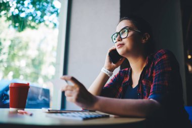 Ekose gömlekli ve gözlüklü ciddi bir kadının modern kafede kahve molası verip gözlerini kaçırırken aramak için akıllı telefon kullanması.