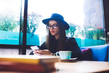 Düşük açılı, şık kıyafetli, bir fincan kahve ve cep telefonuyla masada oturan düşünceli bir kadın serbest yazar.