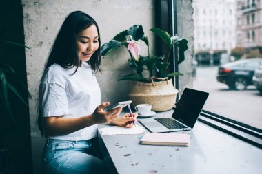 Kafede dizüstü bilgisayarlı, not defterli ve akıllı telefondan mesaj atan gülümseyen Asyalı kız öğrenci.