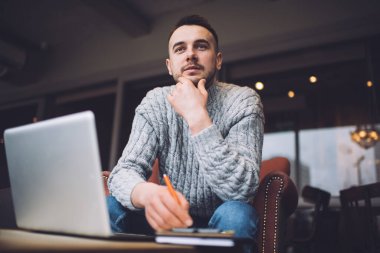 Düşük açılı, eli çenesinde bir adam masanın yanında dizüstü bilgisayarı açık bir şekilde otururken kafasını çeviriyor.