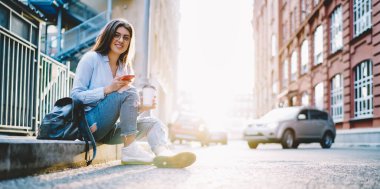  Güler yüzlü hipster blog yazarı, elinde içecek ve akıllı telefon teknolojisiyle güneşli bir havada Kentsel caddede poz veriyor.