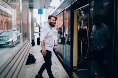 Kol saati takmış, gündüzleri otobüs beklerken kameraya bakan sıradan giyinmiş neşeli sakallı adamın yan görüntüsü.