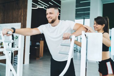 Spor kıyafetleri içinde, spor salonunda spor yaparken sakallı sporcunun kollarını destekleyen, modern spor aletleri üzerinde egzersiz yapan pozitif bir sporcu.