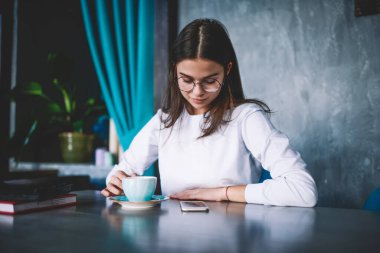 Beyaz elbiseli ve gözlüklü genç bir kadın cep telefonundan mesaj okuyor ve akşam yemekhanedeki masada sıcak içecek içiyor.