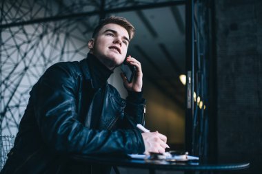 Siyah deri ceketli, balıkçı yakalı ve elinde kalemle akıllı telefondan iletişim kuran yakışıklı bir erkek.