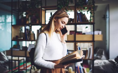 Modern kitapçıda elinde kitapla durup telefonla konuşan ve sıradan kıyafetler giyen genç bir bayanın yan görüntüsü.
